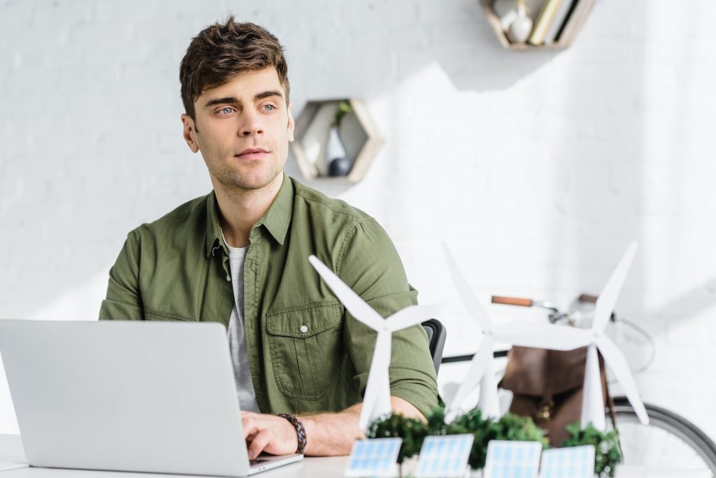 selective focus of handsome architect at table with laptop, solar panels models, windmills and trees